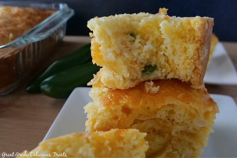Cheesy Cornbread is a great side dish recipe to make for your favorite meals. A white plate with three pieces of cornbread on it, 2 jalapenos, and the baking dish of cornbread in the background.