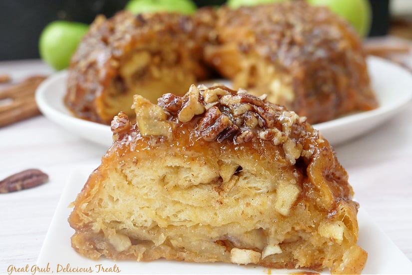 A slice of cinnamon roll on a white plate with the more cinnamon rolls in the background.
