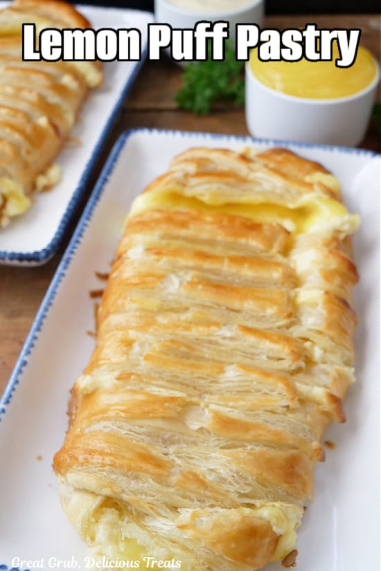 Two lemon puff pastries on two rectangle white plates with blue trim with two small white bowls filled with lemon pie filling and cream cheese mixture.
