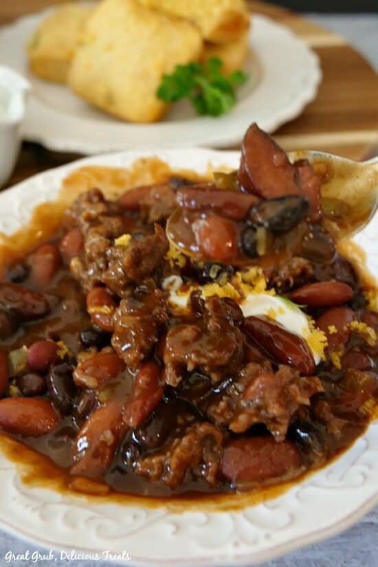 One pot kielbasa chili is a delicious dinner recipe that is full of hearty beans, sausage, and kielbasa. A close up photo of chili in a white bowl topped with sour cream and cheese and a white plate with cornbread stacked.