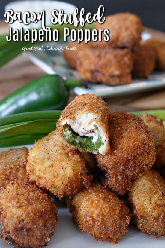 Stuffed jalapeno poppers stacked up on a white plate with a bite taken out of one of them, with green onions and a jalapeno in the background, and another white plate with jalapeno poppers stacked up.