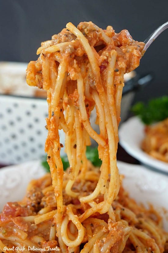 Italian Sausage Spaghetti Bake is a filling and creamy pasta recipe, loaded with Italian sausage, cheese and cream cheese. A forkful of Italian Sausage Spaghetti Bake being held up over a serving bowl of spaghetti bake with another bowl and the white and black polka dot baking dish in the background.